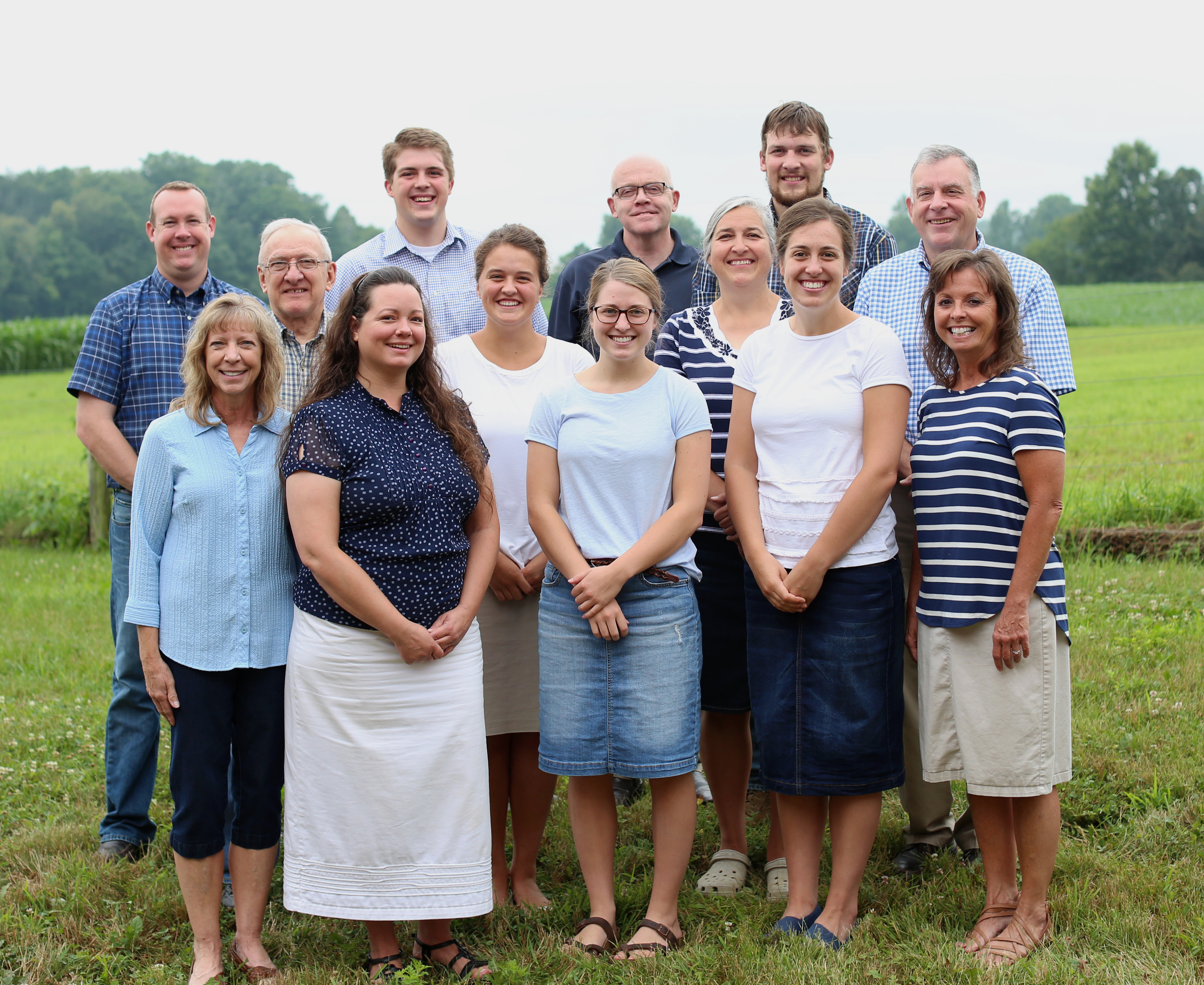 Meet the team at Amish Outlet Store, standing outdoors with a green field in the background. The group consists of smiling men and women, all dressed modestly, representing the dedicated professionals behind the Amish hardwood furniture business.